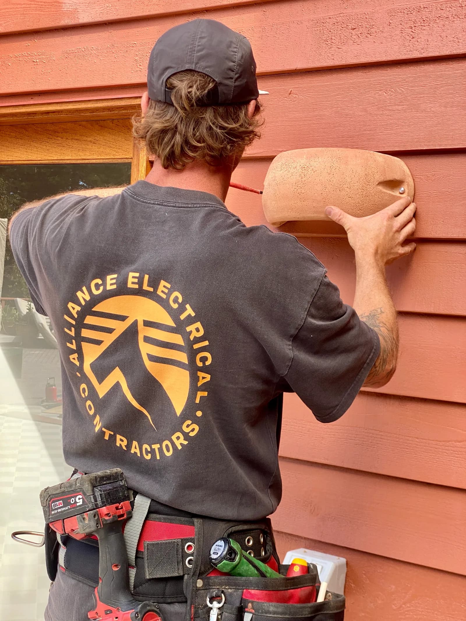 An Alliance Electrical electrician in a branded work shirt fitting a round wall light to the weatherboard wall of a house.