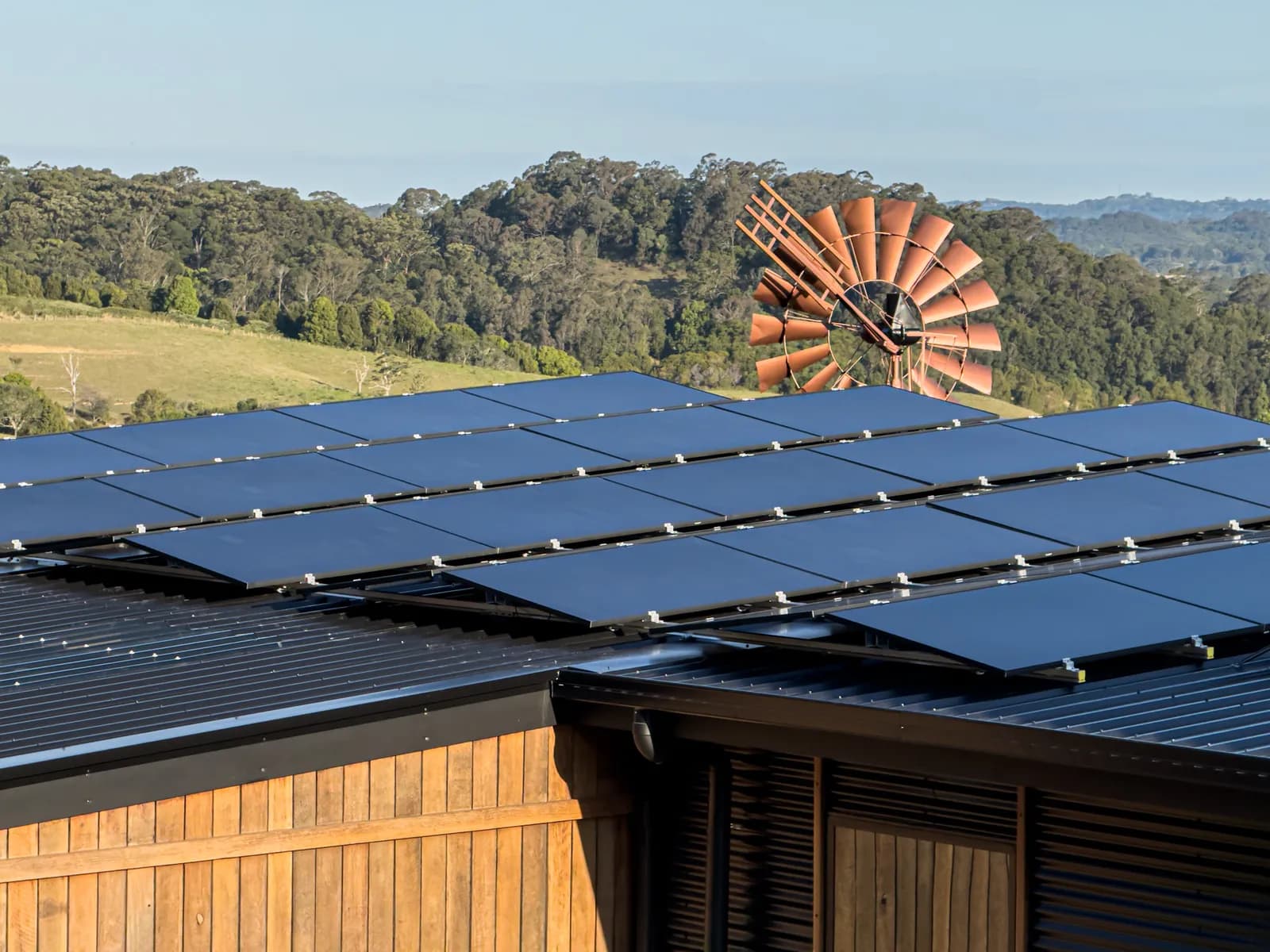Solar panels covering the roof of a timber clad building, with a windmill and green hills behind.